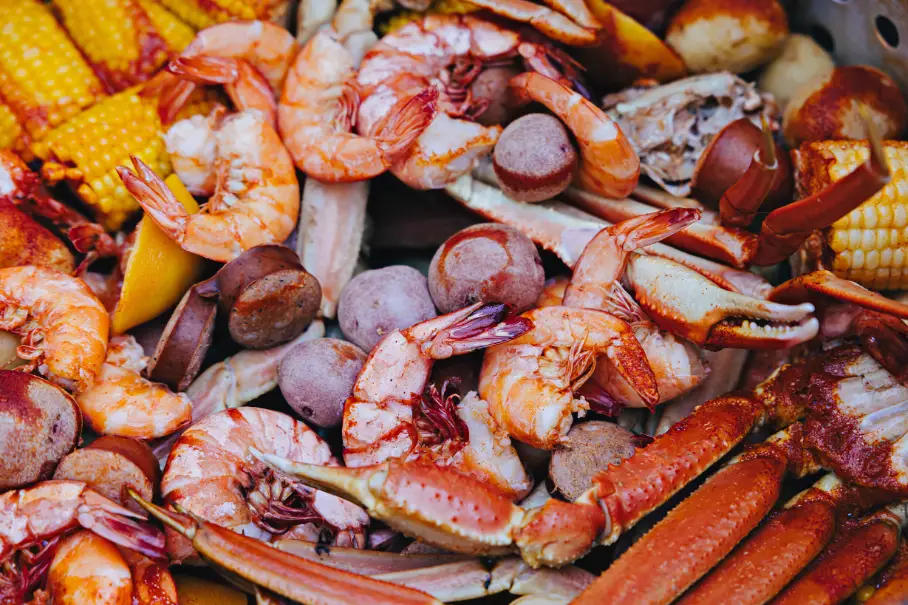 A close up image of a seafood boil. The seafood boil consists of crab legs, shrimp, corn on the cob, and sausage. The crab legs are red and orange in color and are the most prominent element in the image. The shrimp are pink and orange in color and are scattered throughout the image. The corn on the cob is yellow and is visible on the left side of the image. The sausage is brown and is visible on the right side of the image. The background is blurred and not visible.