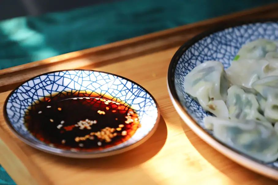 A plate of steamed dumplings and a dish of dipping sauce on a wooden table with a green and blue tablecloth.