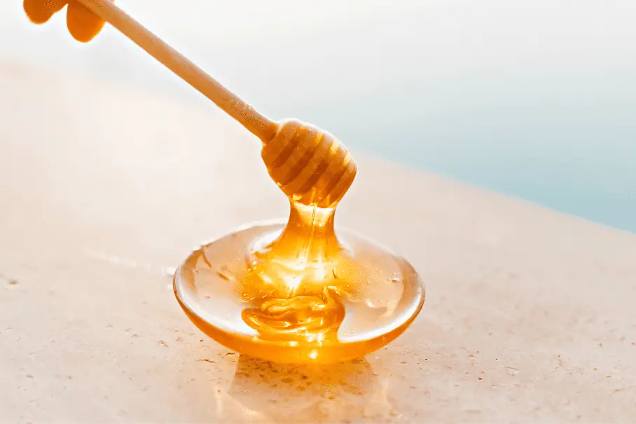 A wooden honey dipper dripping honey onto a plate on a light blue background.