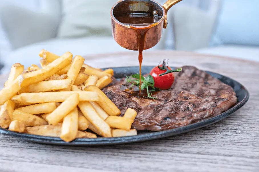 A plate of steak and fries with sauce being poured over the steak on a wooden table.