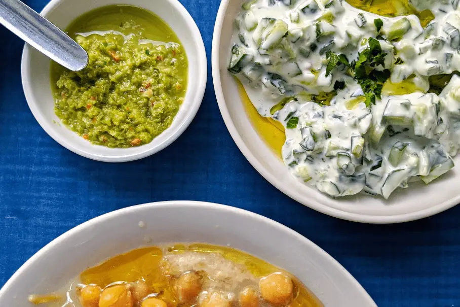 Three bowls of sauce and tzatziki with chickpeas and herbs on a blue tablecloth.