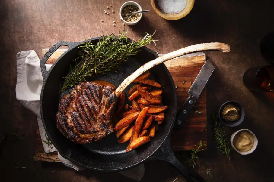 A cast iron skillet with a steak and sweet potato fries on a wooden table, with sprigs of rosemary, salt, pepper, mustard, and a knife on the side. The steak has a bone and grill marks, and the fries are thick wedges. The background is dark and blurry.