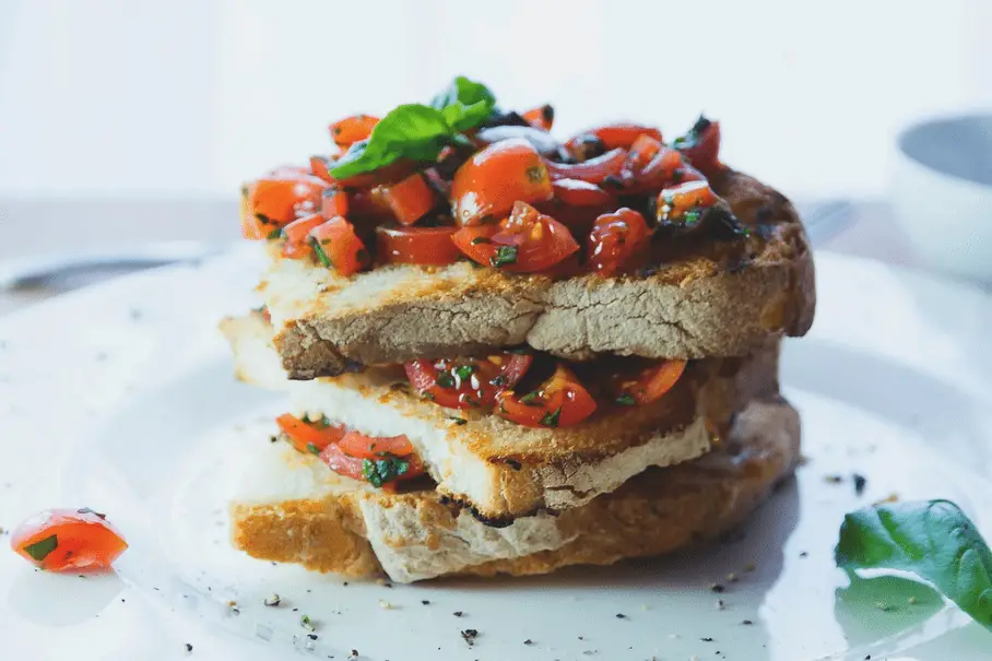 A sandwich with tomatoes on a white plate on a kitchen counter with a bowl and a spoon.