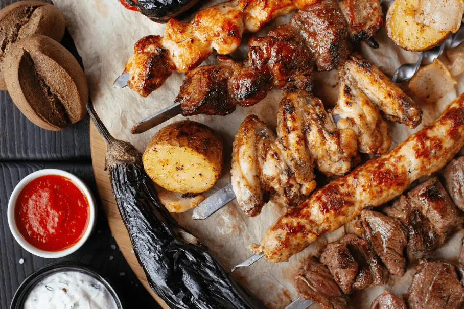A platter of grilled meat and vegetable skewers on a wooden table with sauces and bread.