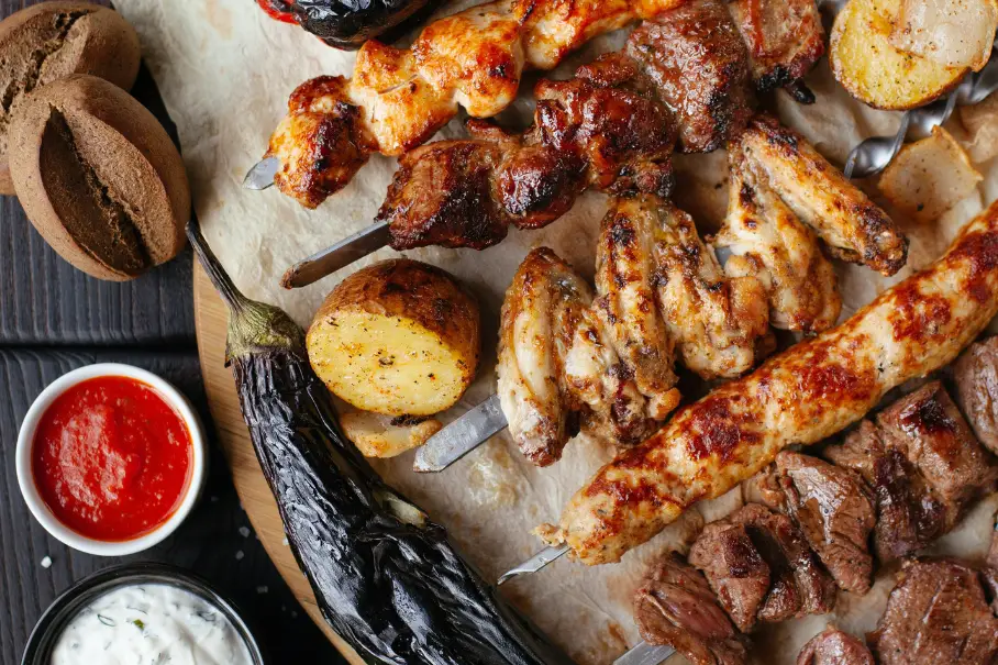 A platter of grilled meat and vegetable skewers on a wooden table with sauces and bread.
