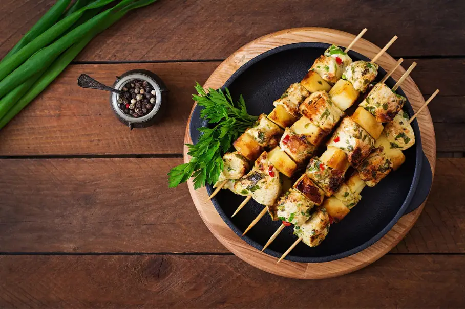 A black plate with grilled paneer skewers on a wooden table with green onions.