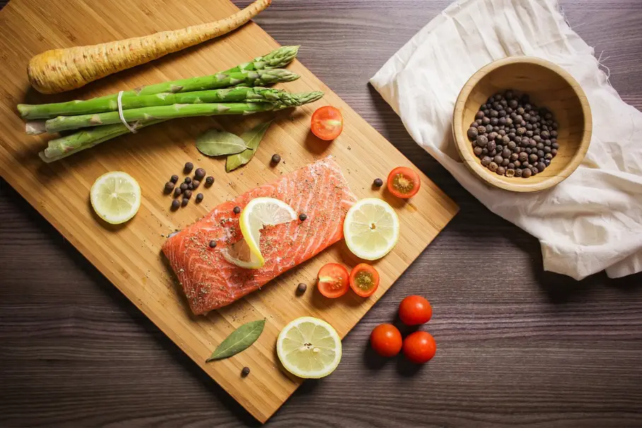 A cutting board with salmon and vegetables on a dark table.