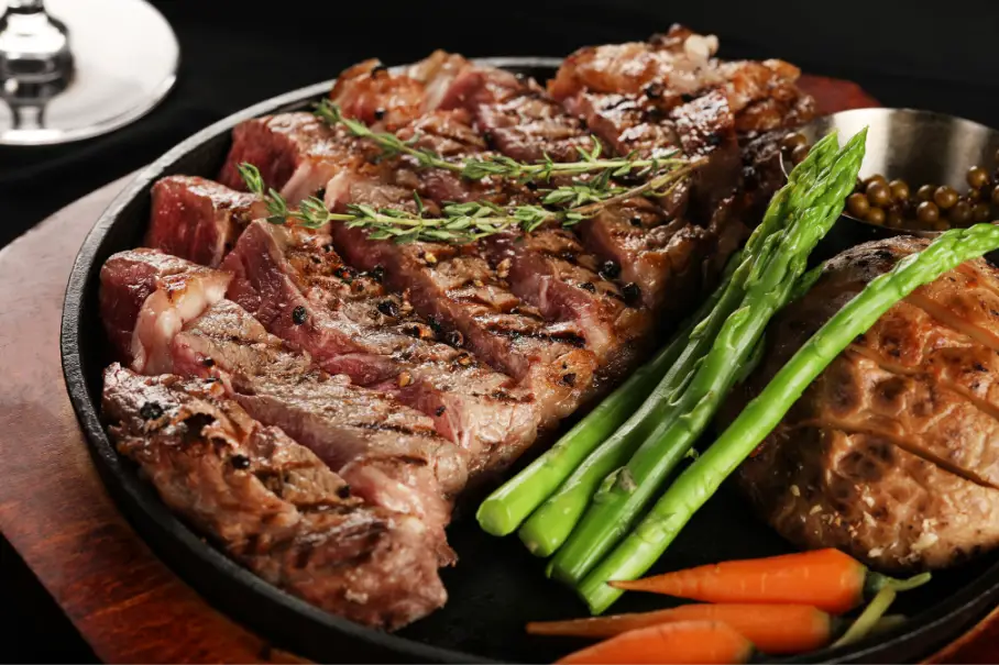A steak dish with rosemary and vegetables on a black plate on a black tablecloth.