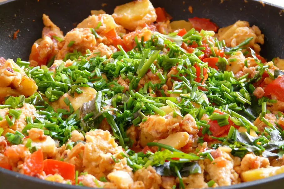 A black frying pan with a stir fry of chicken, red bell peppers, potatoes, and green onions, garnished with chives, on a stovetop.