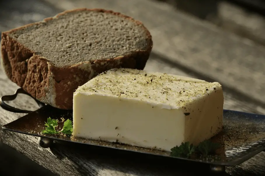 A block of yellow butter with green herbs on a dark plate, with a slice of brown bread next to it, on a wooden table.