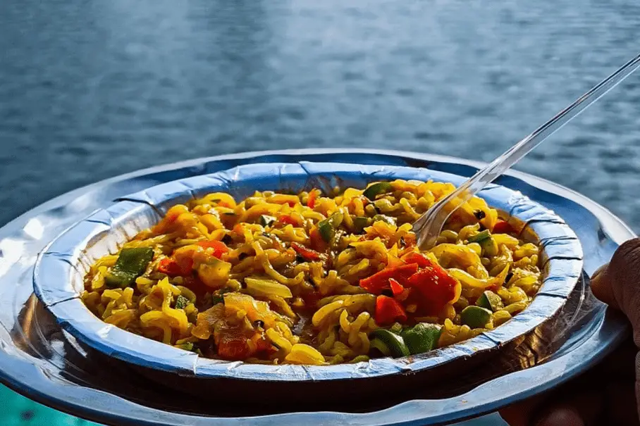 A plate of yellow Maggi noodles with peppers, onions, and vegetables, with a plastic fork in it, on a metal plate, near a body of water.