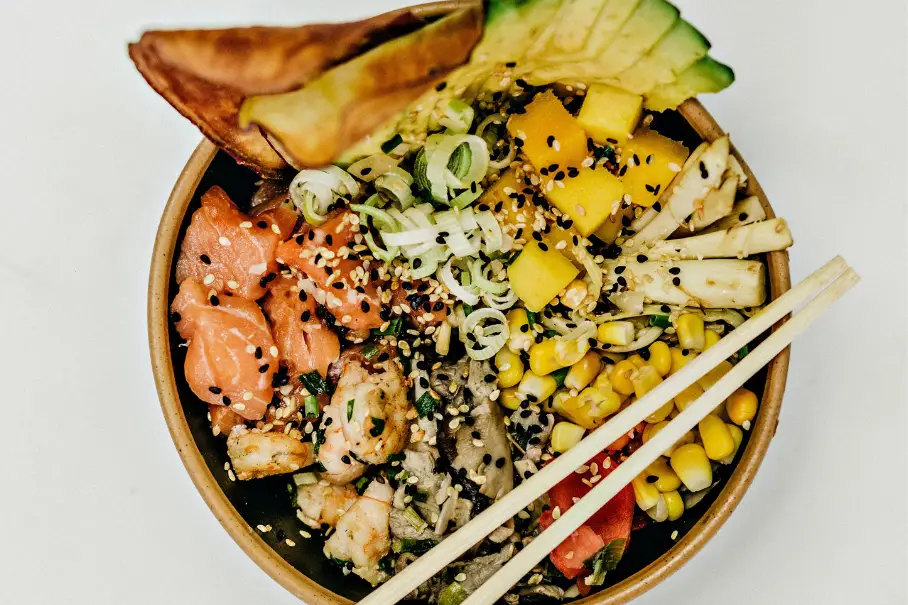 A wooden bowl of poke with salmon, shrimp, avocado, mango, corn, and other toppings, with chopsticks on a white background.