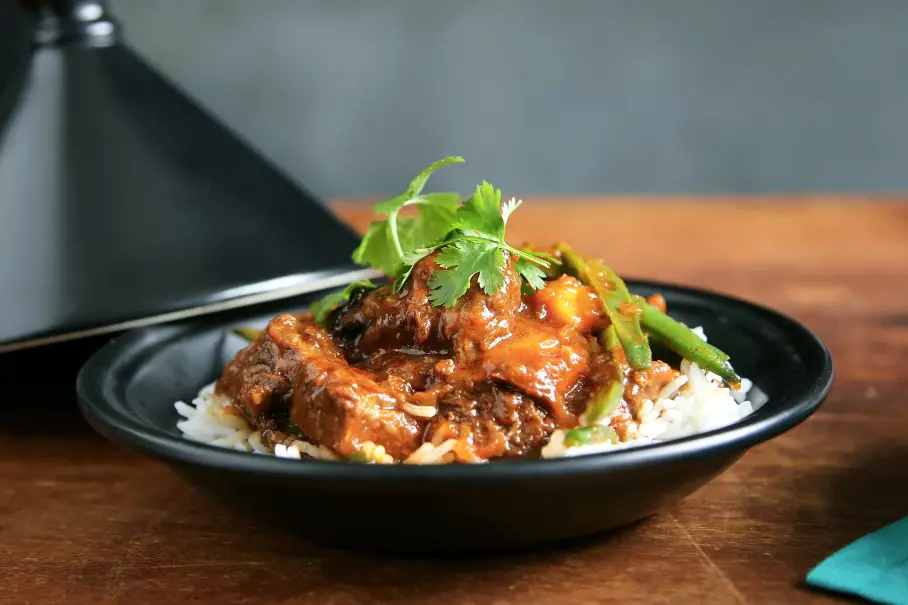 A black bowl of beef and rice dish with cilantro and green peppers on a wooden table. The image has a blurred background with a black lamp.