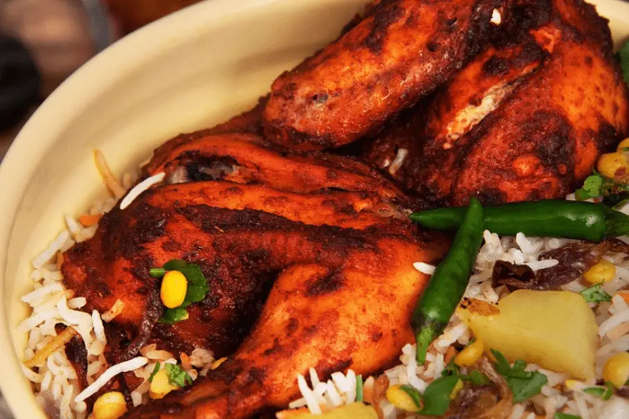 A photo realistic image of rice and grilled chicken. The chicken is marinated in spices and has a dark red color. The rice is mixed with peas, corn, and diced potatoes. There is a green chili pepper on top of the dish. The background is blurred and appears to be a restaurant setting.