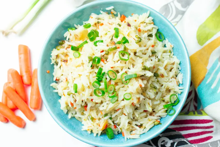 A photo realistic image of a blue bowl of fried rice with green onions and carrots on a white background. The bowl has a white rim and is filled with white rice with bits of orange and red. The green onions are sliced and scattered on top of the rice. The carrots are whole and are on the left side of the image. The background is white with a striped cloth on the right side of the image.