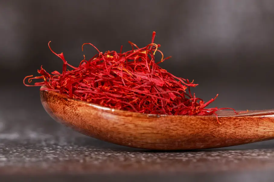A photo realistic image of a wooden spoon filled with red saffron threads on a dark grey background. The saffron threads are bright red and appear to be fresh. The wooden spoon is a dark brown color and has a smooth texture. The background is a dark grey color with a slight texture.