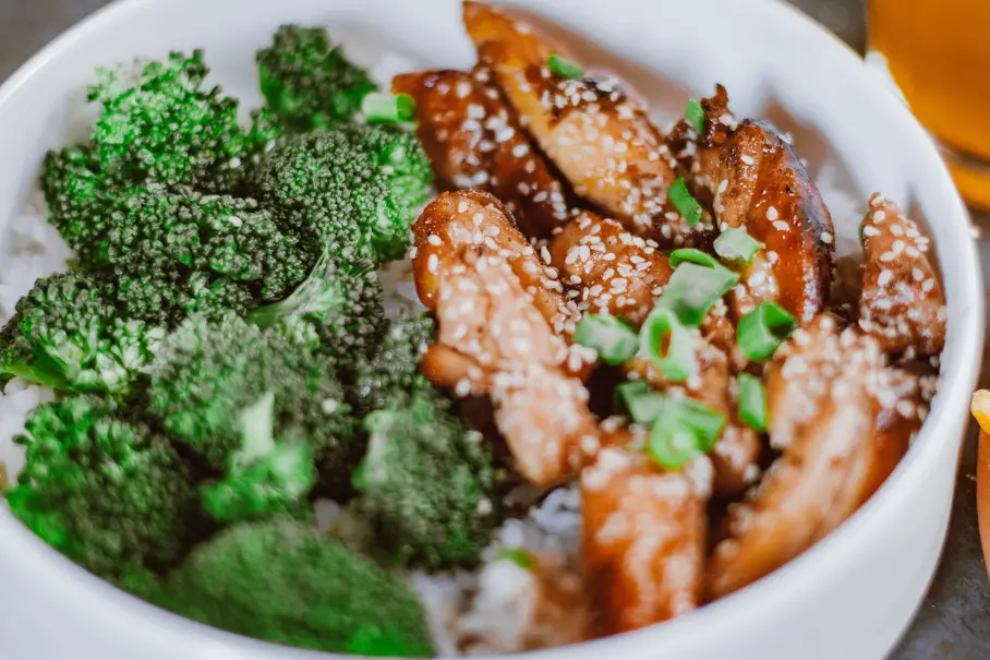 A photo realistic image of a white bowl of food consisting of broccoli and chicken. The broccoli is bright green and appears to be steamed. The chicken is covered in a dark sauce and sesame seeds. The chicken is sliced and arranged in a way that it covers half of the bowl. The bowl is on a wooden surface and there is a blurred glass in the background.