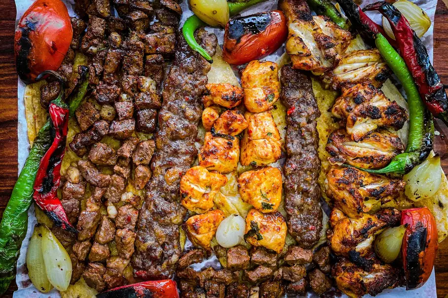 A photo realistic image of a large platter of grilled meat and vegetables. The platter is rectangular and made of wood. The meat is in the center of the platter and is a mix of beef and chicken. The beef is cubed and the chicken is in the form of kebabs. The vegetables are a mix of bell peppers, onions, and tomatoes. The vegetables are grilled and charred. The platter is garnished with parsley and lemon wedges. The background is a wooden table.