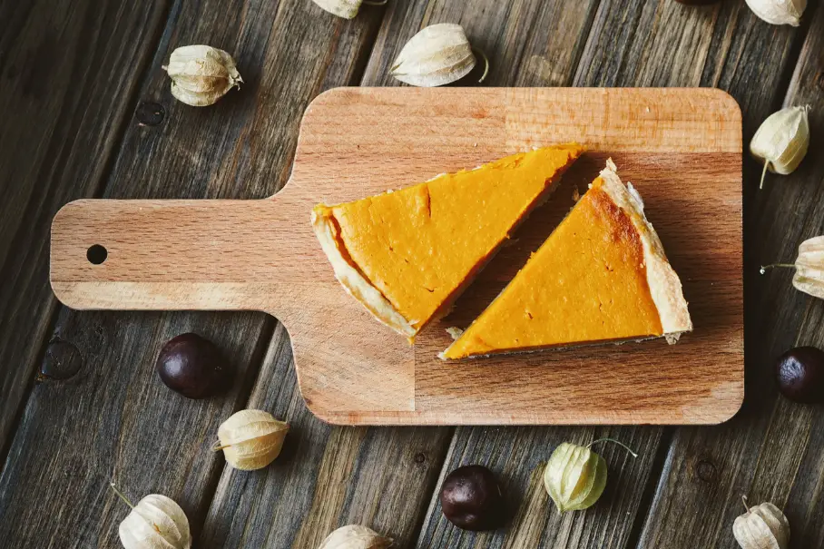 A photo realistic image of two slices of sweet potato pie on a wooden cutting board. The cutting board is rectangular with a handle on one end. The pie slices are triangular and have a golden brown crust. The filling is a smooth orange color. There are scattered chestnuts and gooseberries around the cutting board. The background is a wooden table with a rustic texture.