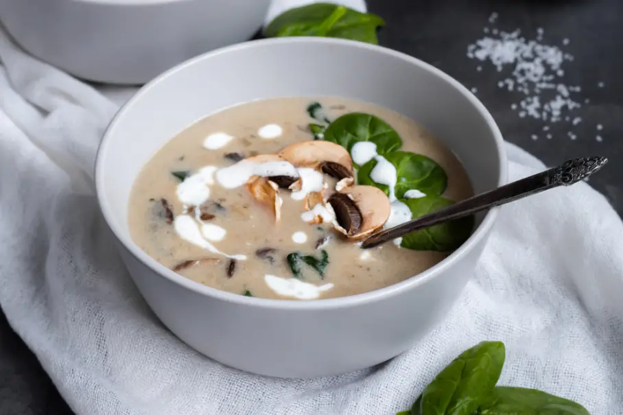 A photo realistic image of a bowl of creamy mushroom soup with a spoon in it. The soup is in a white bowl with a silver spoon. The soup is garnished with sliced mushrooms, fresh basil leaves and a drizzle of cream. The bowl is on a white cloth napkin with a sprinkle of salt on the side. The background is a dark grey countertop.