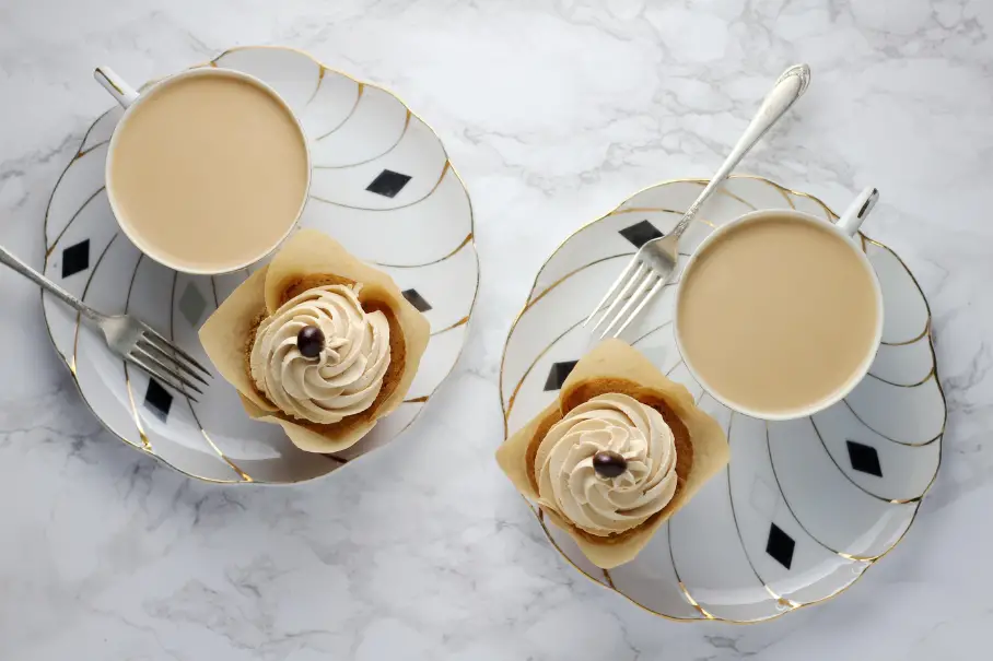A photo realistic image of two cups of tea and two slices of cake on a marble surface. The cups and saucers are white with a gold and black geometric pattern. The tea is a light brown color and the cups are filled to the brim. The cake is a light brown color with a swirl of white frosting on top and a single chocolate chip in the center. The forks are silver and are resting on the saucers. The background is a white marble surface with gray veining.