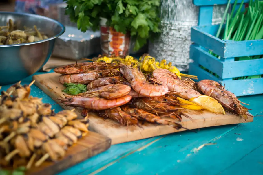 A cutting board with grilled shrimp on a blue table with skewers of meat and vegetables and bowls of food in the background.