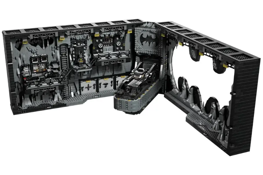 An image of a LEGO set of the interior of the Batcave from the Batman series. The set is made up of black and gray LEGO bricks. The set is in an L shape with the left side being the main part of the Batcave and the right side being the entrance. The left side of the set has a computer console, a batmobile, and a batwing. The right side of the set has a batcycle and a batboat. The set has a lot of details such as pipes, gears, and computer screens. The set is on a white background.