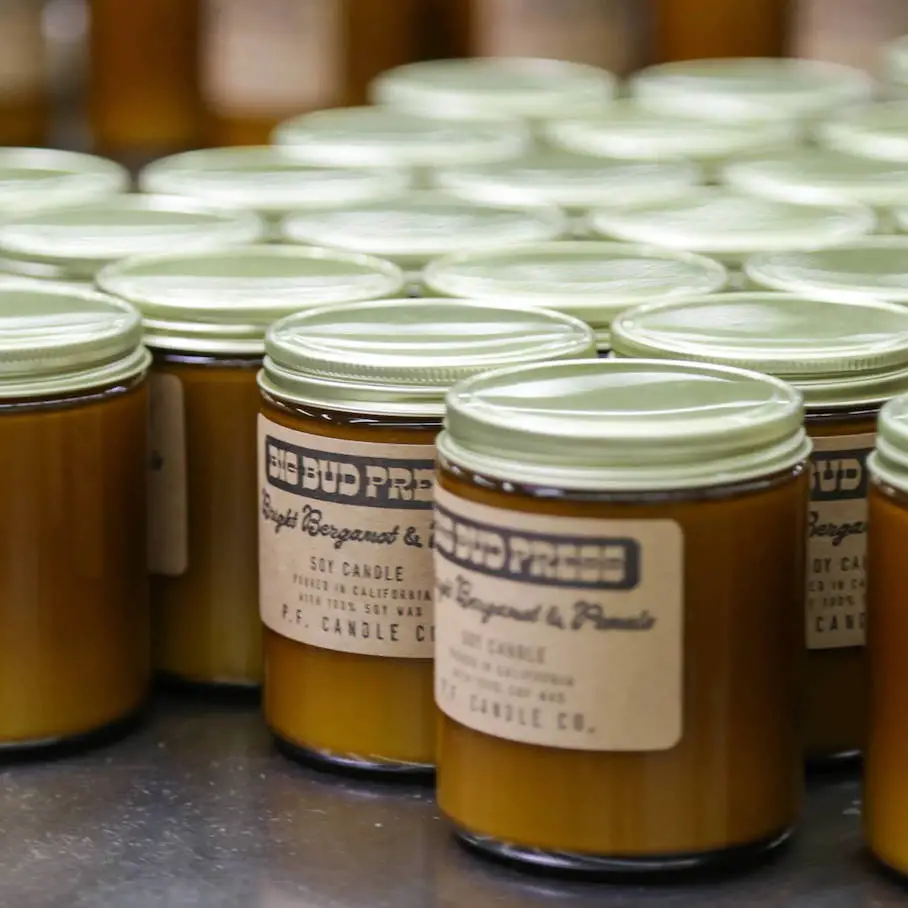 A photo realistic image of rows of small jars with golden lids. The jars are filled with a light brown substance. The jars have a label on them that reads “Big Bud Press” and “Soy Candle Co.”. The jars are arranged in rows on a white surface. The background is blurred and consists of more jars and bottles.