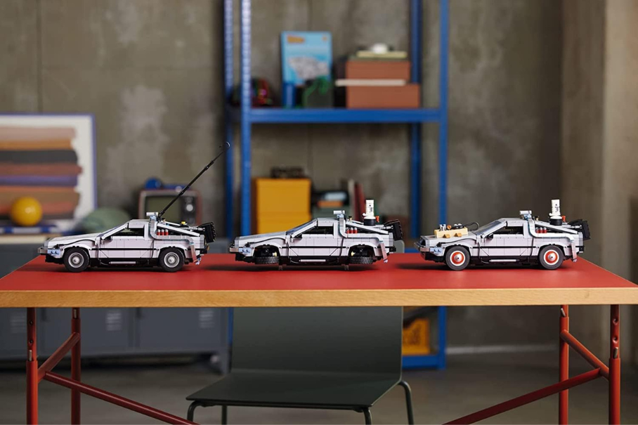 Three toy models of the DeLorean car from the movie “Back to the Future” are displayed on a red table. The models show different stages of modification, from the original car to the fully equipped time machine. The background is a concrete wall and a blue shelf with various items.