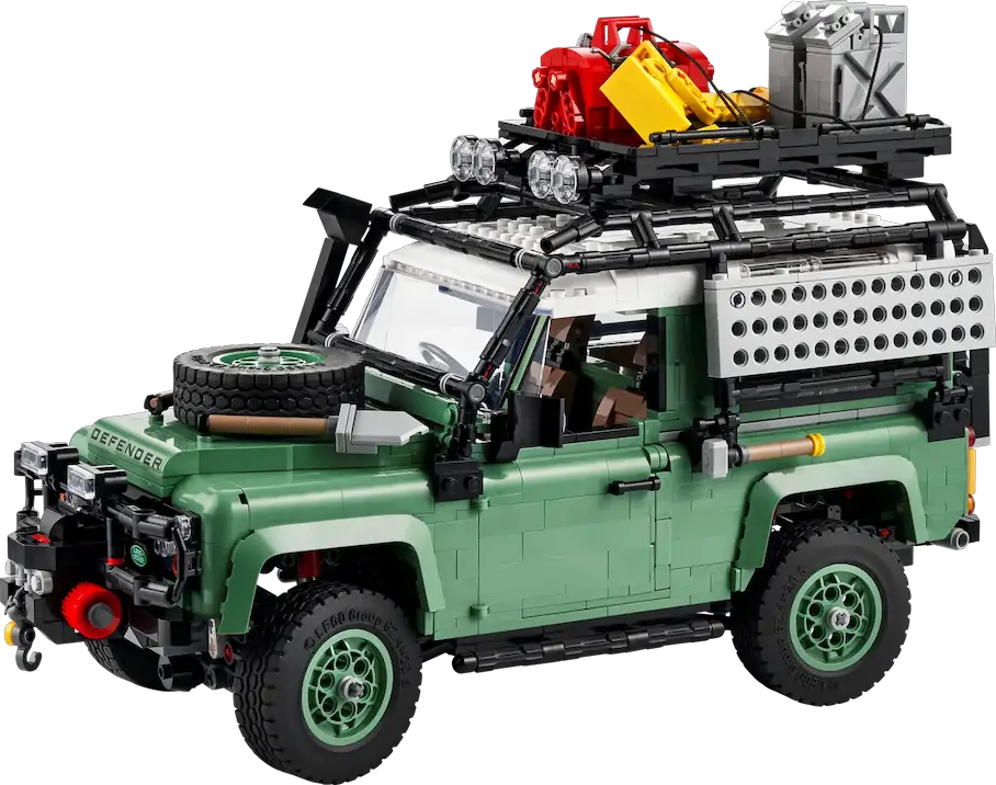A green Land Rover Defender with a black roof rack and a spare tire on the hood. The roof rack has a red and yellow excavator and a gray crate on it. The car has a license plate that reads 'DEFENDER'. The car has black and red accents and a black grill. The background is white.