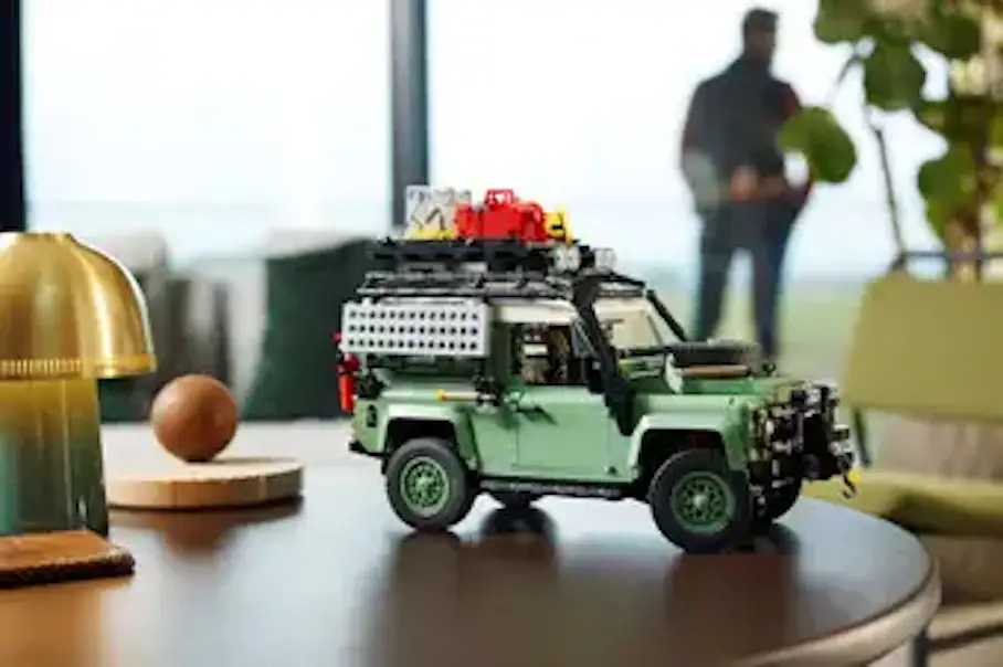 A miniature toy car on a wooden table. The car is a green Land Rover Defender with a black roof rack and red and yellow accessories. The car is parked on a wooden table with a gold lamp and a wooden sphere in the background. The background consists of a blurred figure of a person and a plant.