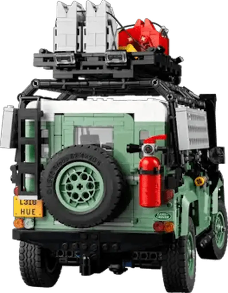 A Lego model of a green Land Rover Defender on a white background. The model has a spare tire on the back and a roof rack with various accessories. The accessories include a red fire extinguisher, a red gas can, and white storage containers.