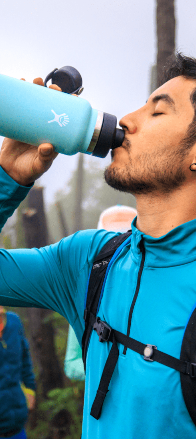 A photo of a person drinking from a blue water bottle. The person is wearing a blue jacket and has a backpack on. The background is a foggy forest with trees and other people visible.