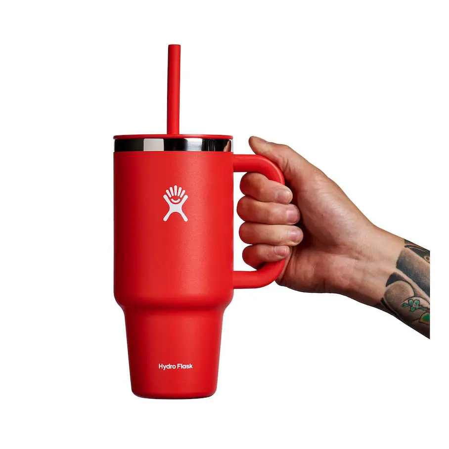 A photo realistic image of a red Hydro Flask tumbler with a straw lid. The tumbler is being held by a hand with tattoos on the wrist. The tumbler has a white logo of a hand with fingers spread out on the side. The straw lid is black and the straw is red. The background is white.