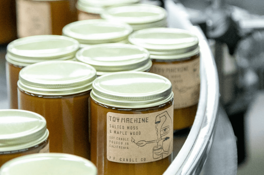 A photo of a group of amber-colored glass jars with white metal lids. The jars have labels on them, one of which reads “TOY MACHINE Salted Moss & Maple Wood 16oz Candle Made in California P.F. Candle Co.”. The jars are arranged in a curved line on a white surface. The background is blurred and appears to be a factory or production line.