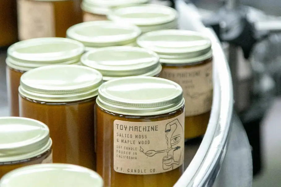 A photo of a group of amber-colored glass jars with white metal lids. The jars have labels on them, one of which reads “TOY MACHINE Salted Moss & Maple Wood 16oz Candle Made in California P.F. Candle Co.”. The jars are arranged in a curved line on a white surface. The background is blurred and appears to be a factory or production line.