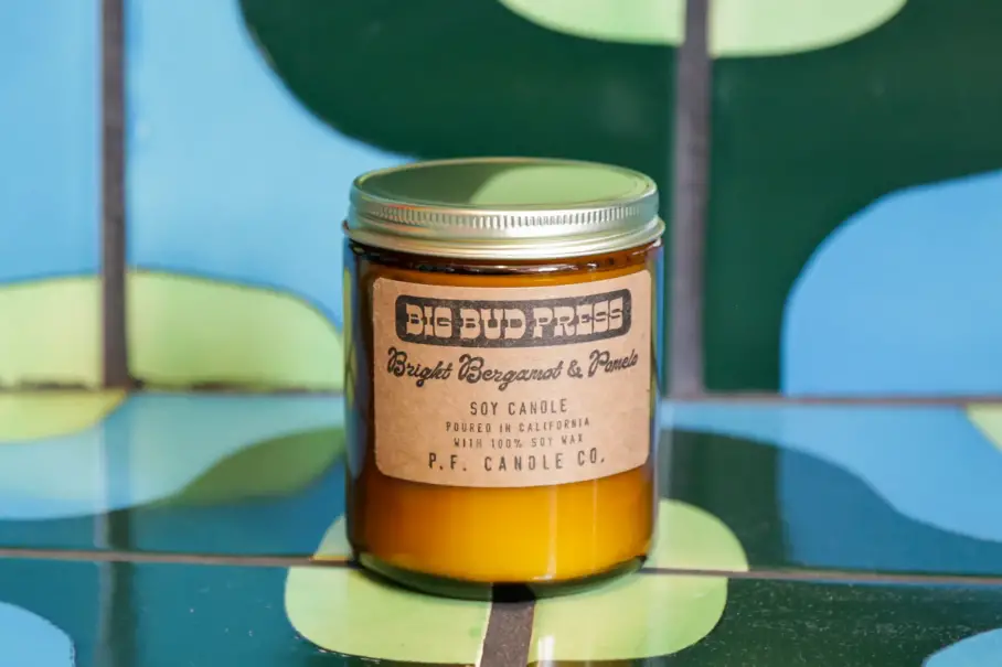 A photo realistic image of a jar of soy candle on a green and blue tiled background. The jar is amber in color with a gold lid. The label on the jar is black with white text. It reads “BIG BUD PRESS Bright Citrus & Basil Soy Candle Poured in California P.F. Candle Co.” The background consists of green and blue tiles with a geometric pattern.
