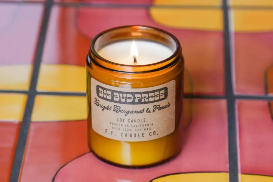 A photo realistic image of a lit soy candle in a small amber jar. The label on the jar is a white rectangle with black text. It reads “Big Bud Press, Bright Bergamot & Basil, Soy Candle, Hand Poured in California, P.F. Candle Co.”. The candle is on a pink and orange tiled surface. The background consists of more of the tiled surface.